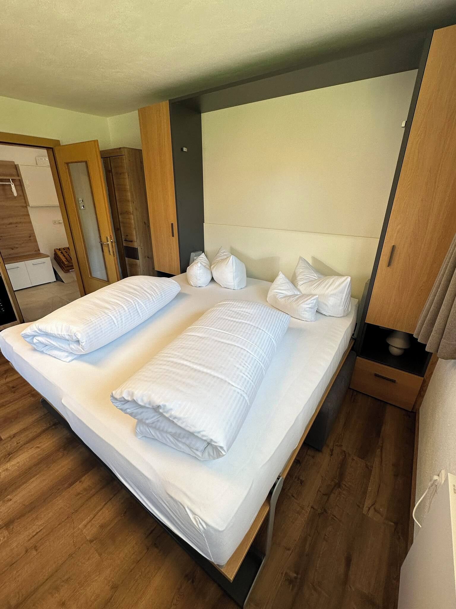 A double bed with white linens and pillows, a wooden headboard, and a wooden floor.