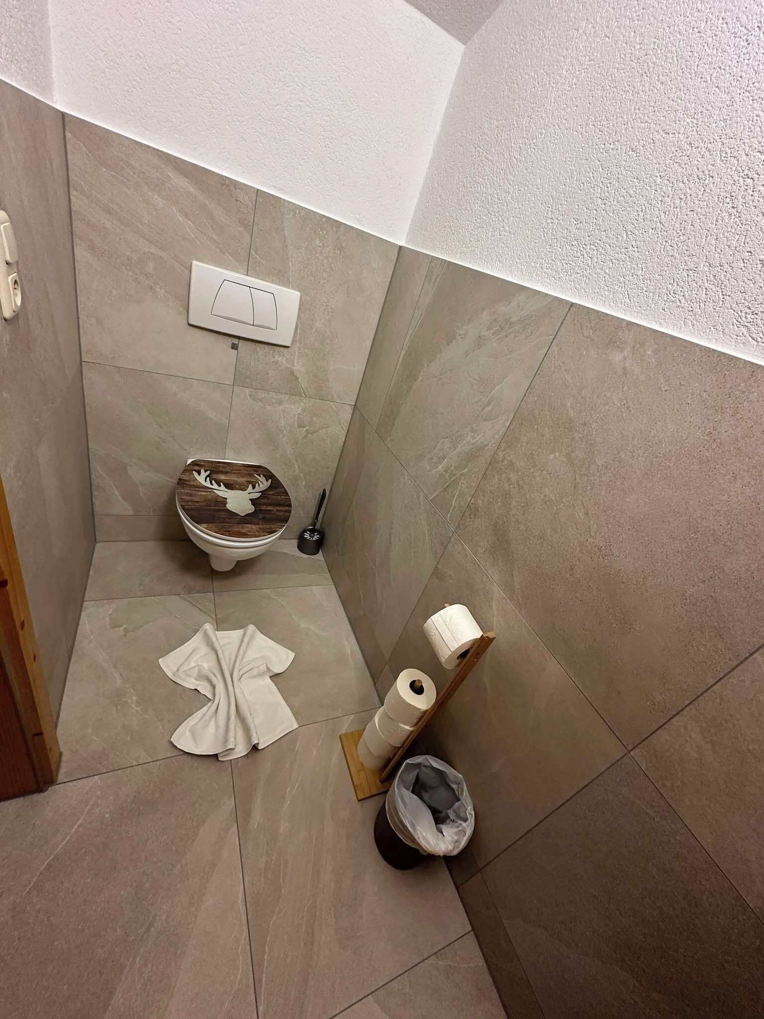 A bathroom with a toilet, a toilet brush, a roll of toilet paper, and a trash bin.