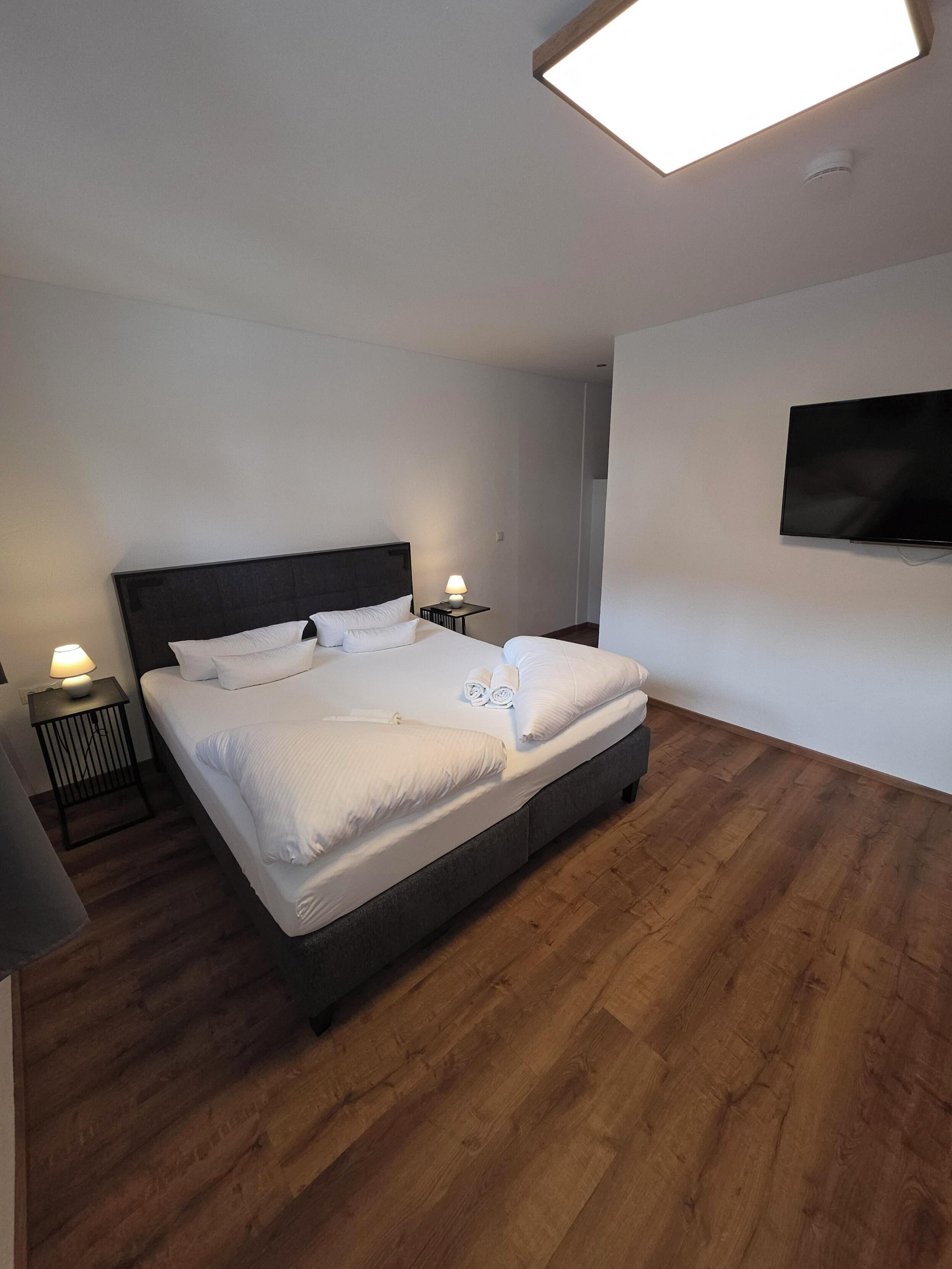 Moderni hotellihuone, jossa on suuri sänky, kaksi yöpöytää valaisimineen, seinälle asennettu litteänäyttöinen televisio ja puulattia.