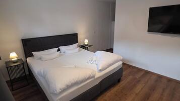 Moderni hotellihuone, jossa on suuri sänky, kaksi yöpöytää valaisimineen, seinälle asennettu litteänäyttöinen televisio ja puulattia.