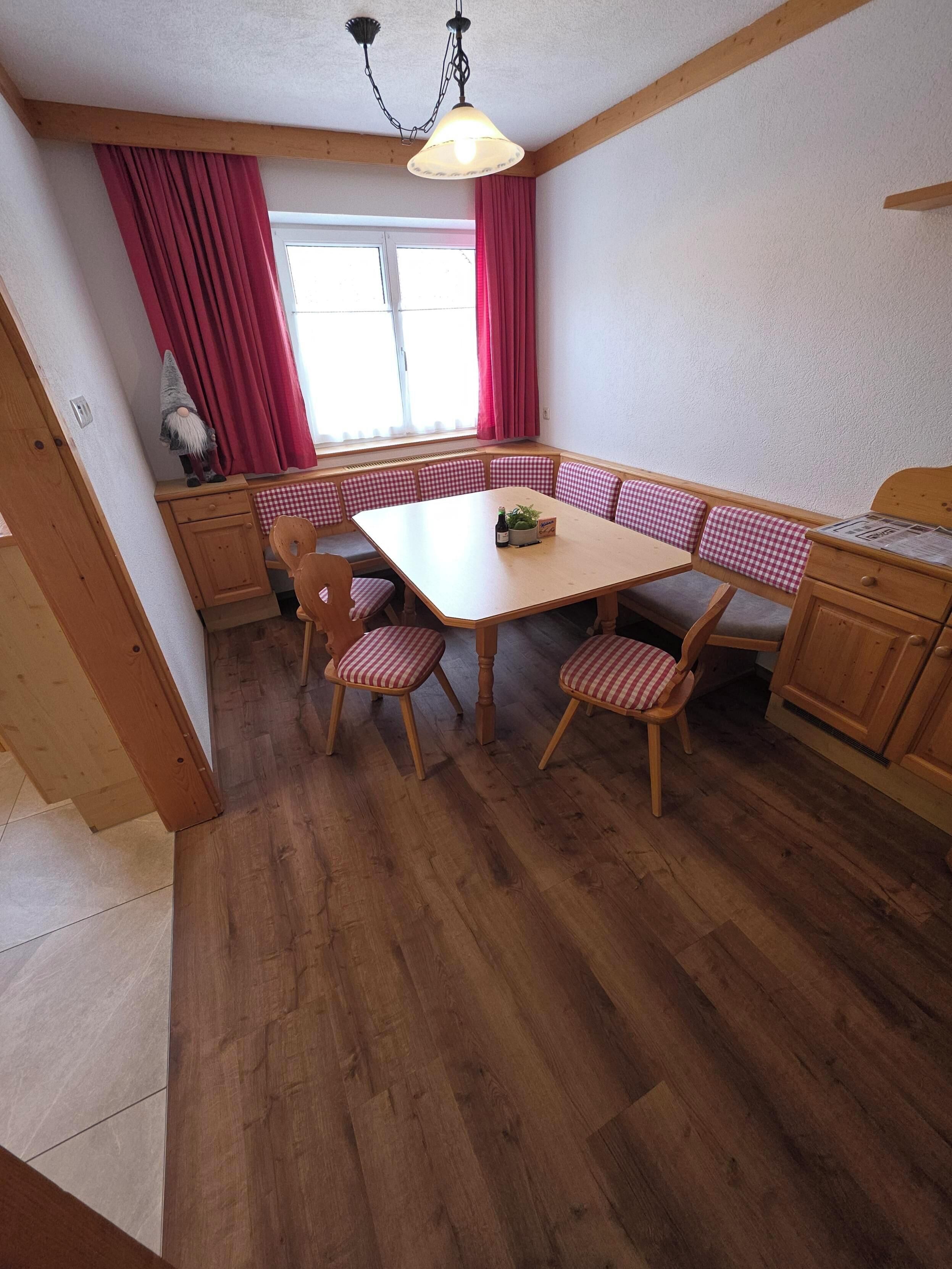 A dining area with a wooden table and checkered chairs, a window with red curtains, and a gnome decoration.