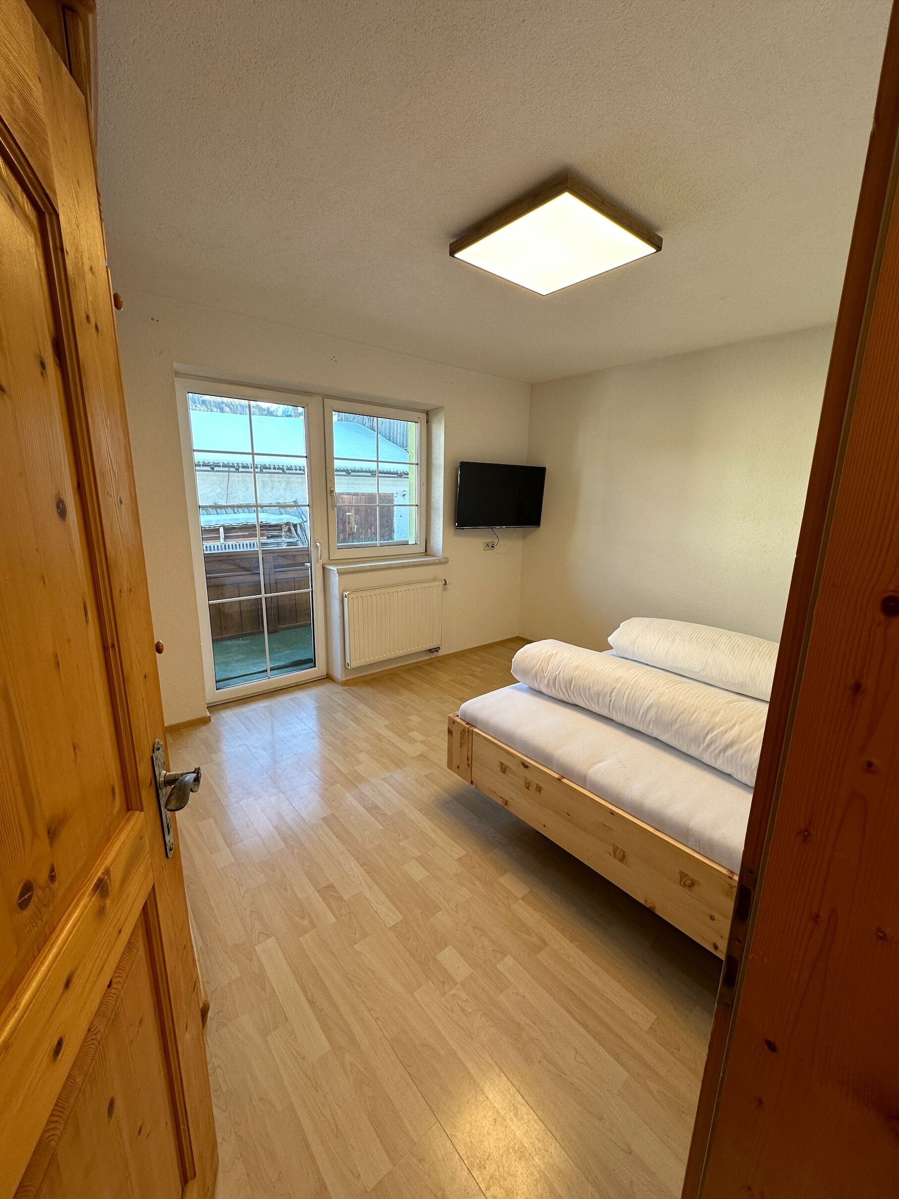 A bedroom with a wooden bed, a window, a radiator, and a TV mounted on the wall.