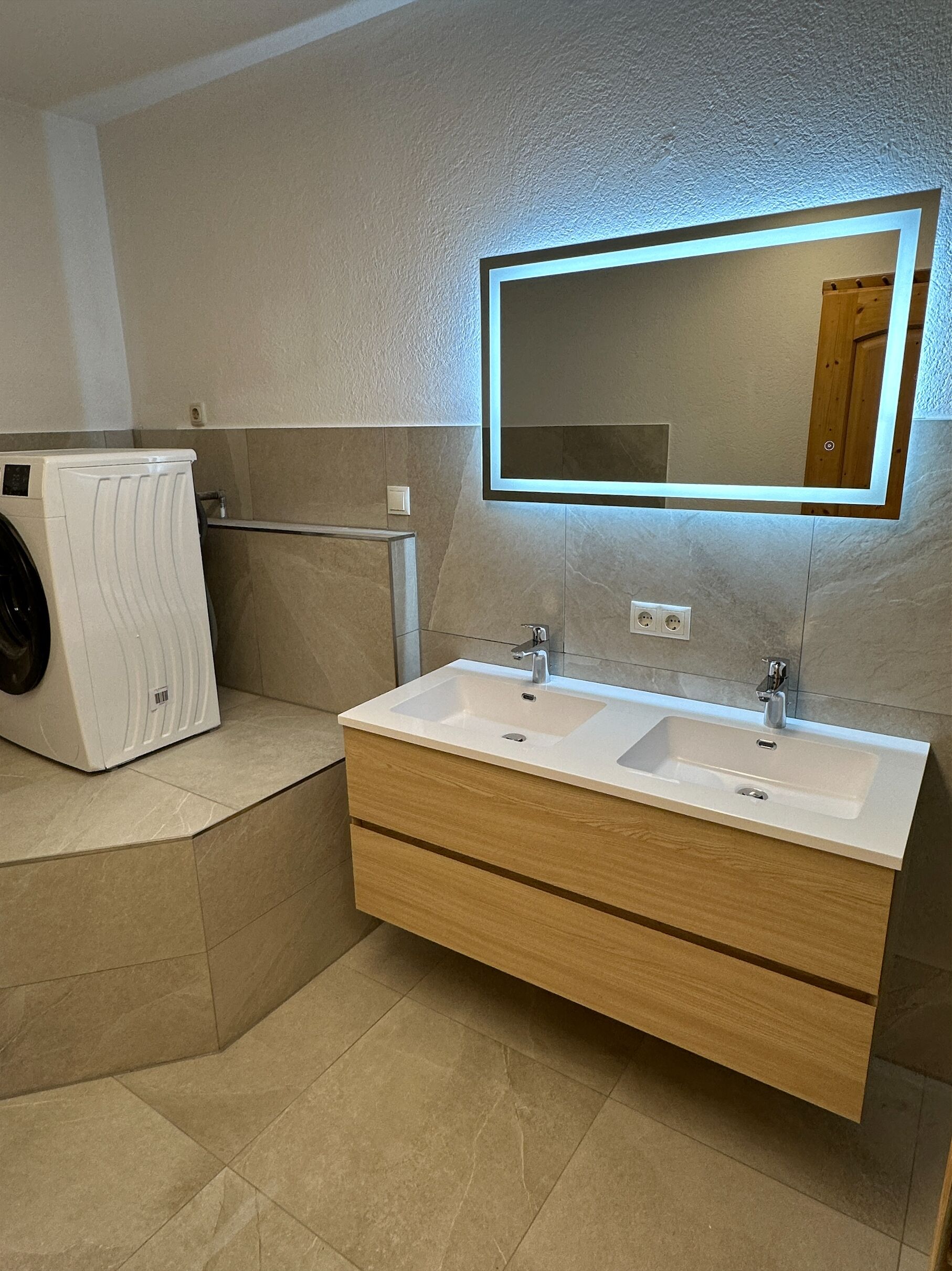 A modern bathroom with a double sink vanity, a built-in washing machine, and a large mirror with LED lighting.