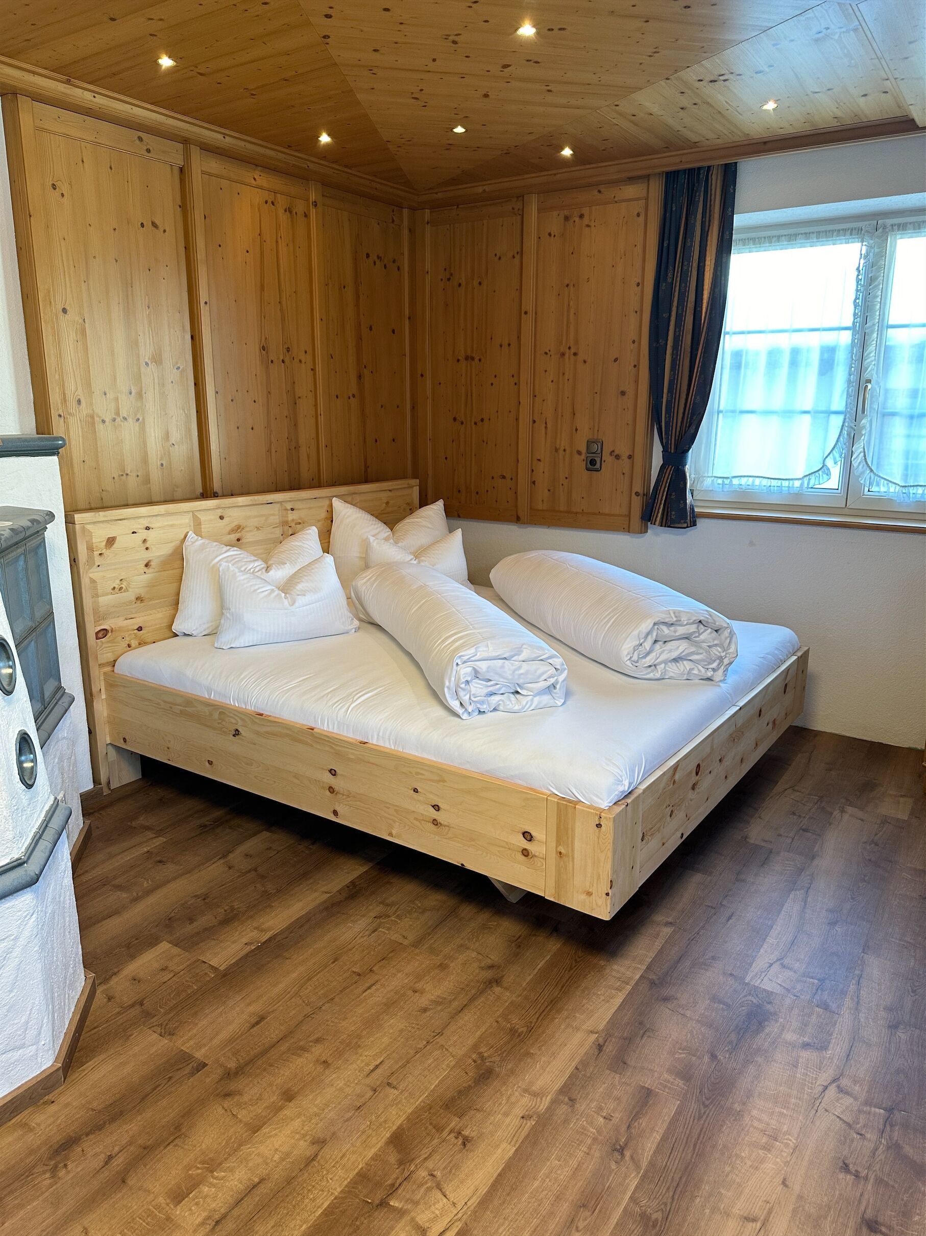 A wooden-paneled room with a single bed, a window with curtains, and a wooden floor.