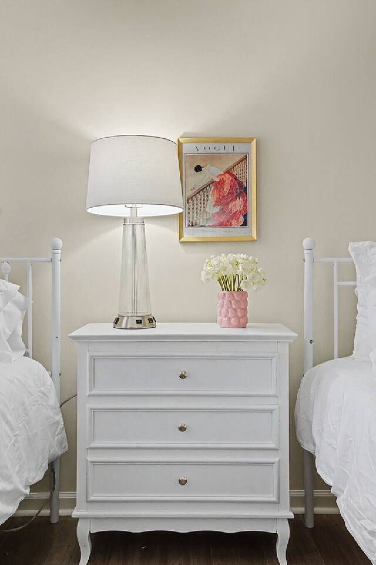A white bedside table with a lamp, a framed picture, and a vase with flowers.