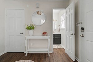 A hallway with a white door, a white console table with a vase of flowers, a round mirror, and a dark wooden floor. - Beautiful Home off Acton Road, 5 Minutes to Summit & 280 (Birmingham)
