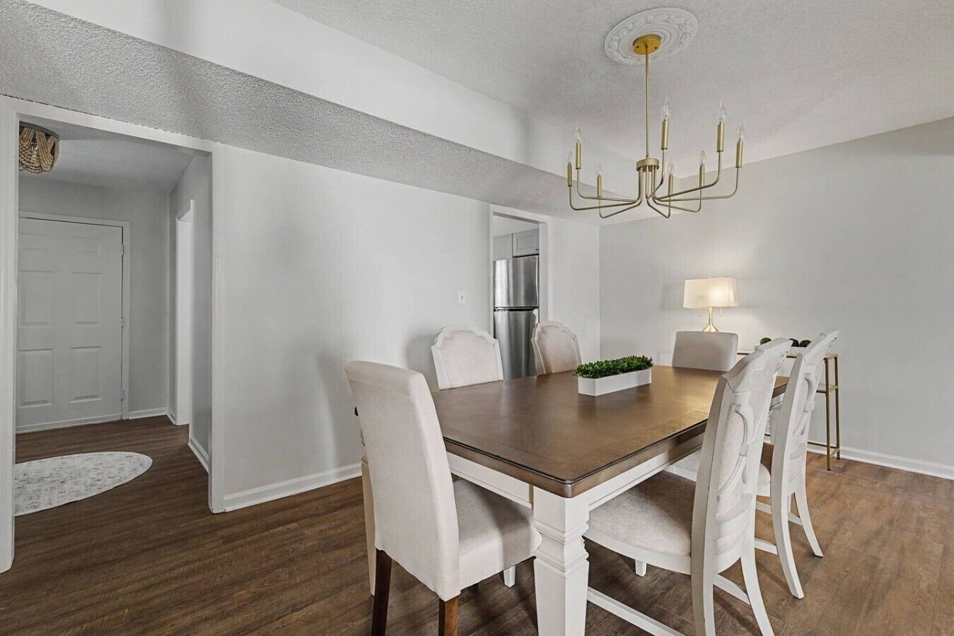 A dining area with a wooden table, white chairs, a chandelier, and a refrigerator in the background.