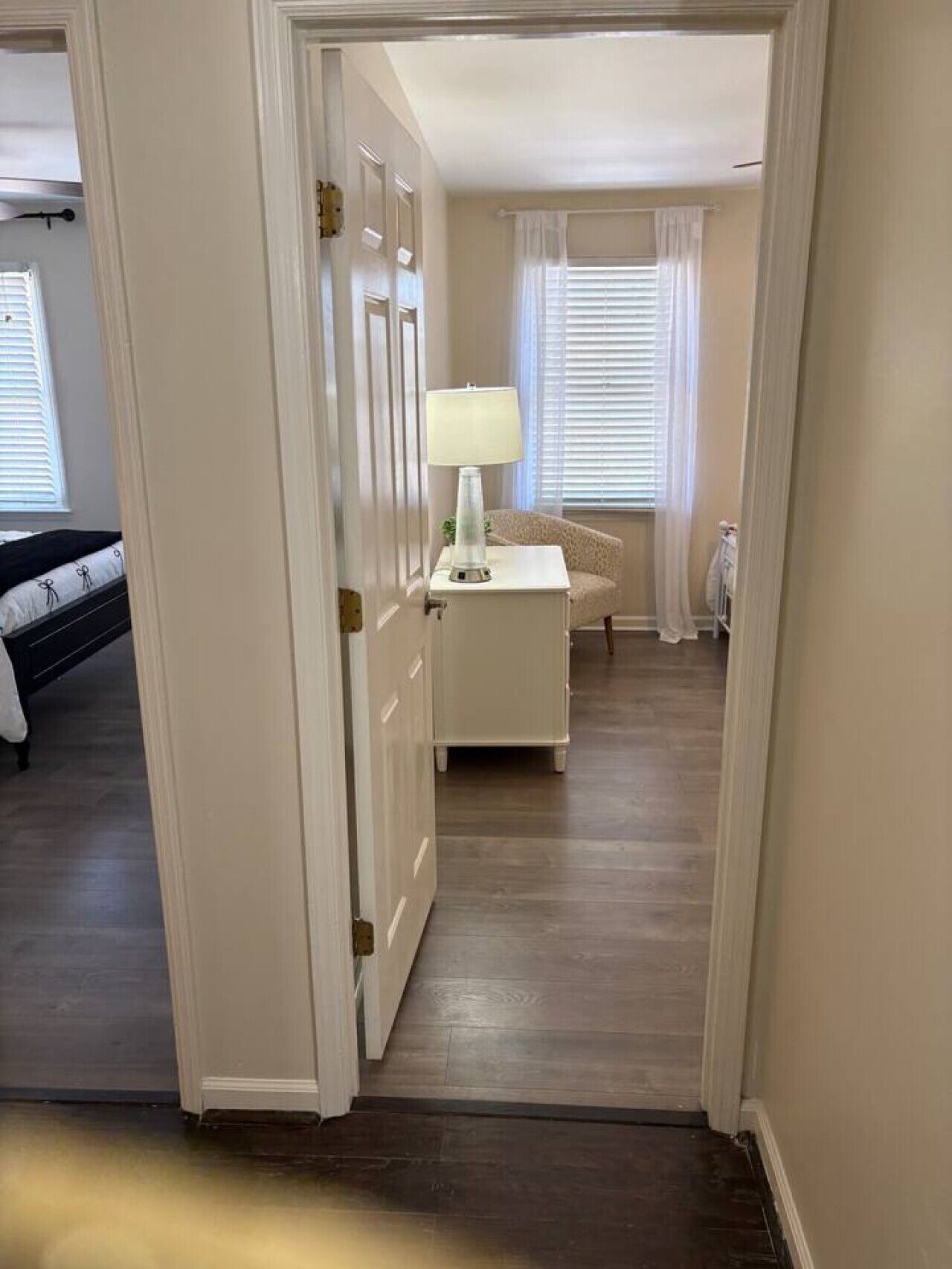 A hallway leading to a bedroom with a bed, a bedside table with a lamp, and a window with blinds.