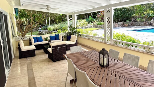 Un espace extérieur aménagé, abrité et doté de sièges, avec vue sur la piscine, comprenant une table en bois et des canapés en osier avec des coussins.