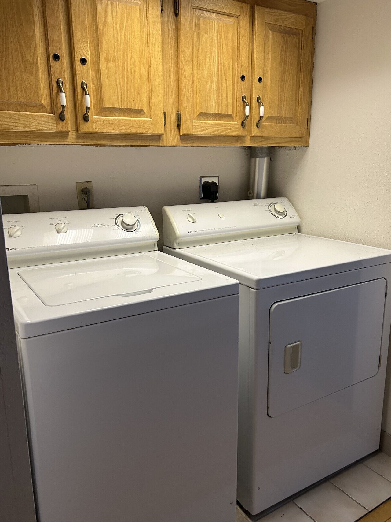 A laundry room with two white front-loading washers and dryers, wooden cabinets above, and a visible electrical outlet.
