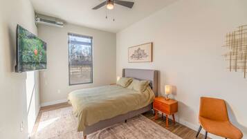 A bedroom with a bed, a nightstand, a chair, a ceiling fan, a window, and a wall-mounted artwork.