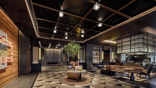 Eine moderne Lobby mit geometrischem Bodenmuster, einem runden Tisch in der Mitte, Sitzbereichen, einem großen Baum und einer Bar.