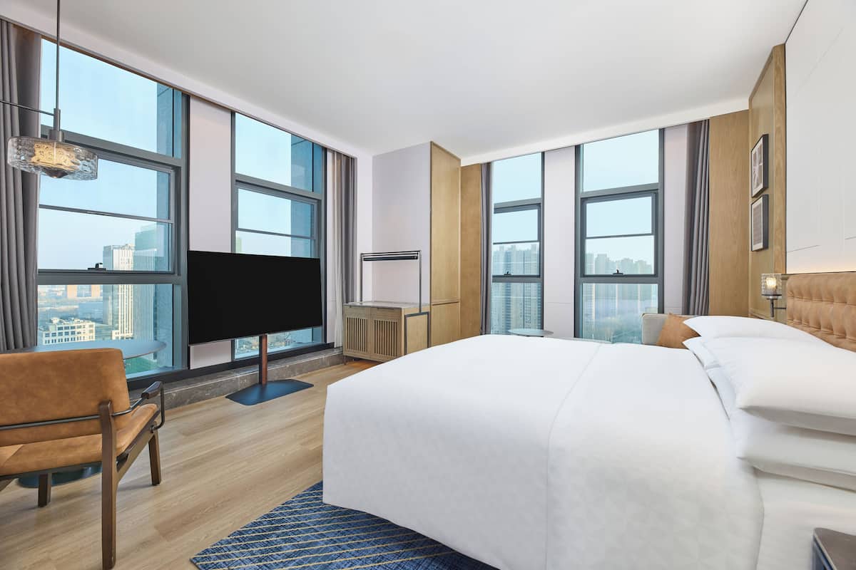 A modern hotel room with a large bed, a flat-screen TV, a seating area, and large windows offering a city view.