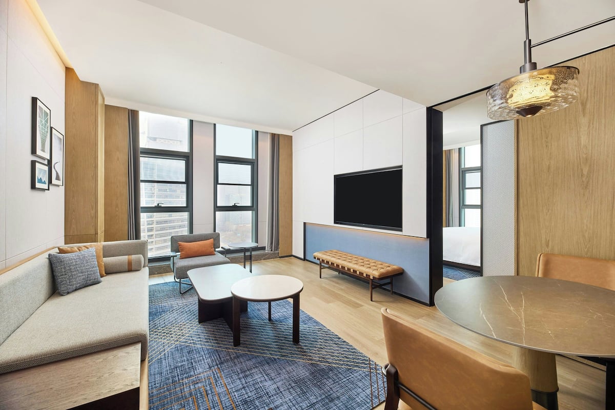 A modern hotel room with a sofa, a round table, a bench, a TV, and large windows.