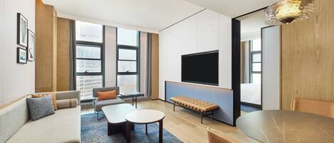 Quarto de hotel moderno com sofá, mesa redonda, um banco, televisão e janelas amplas.