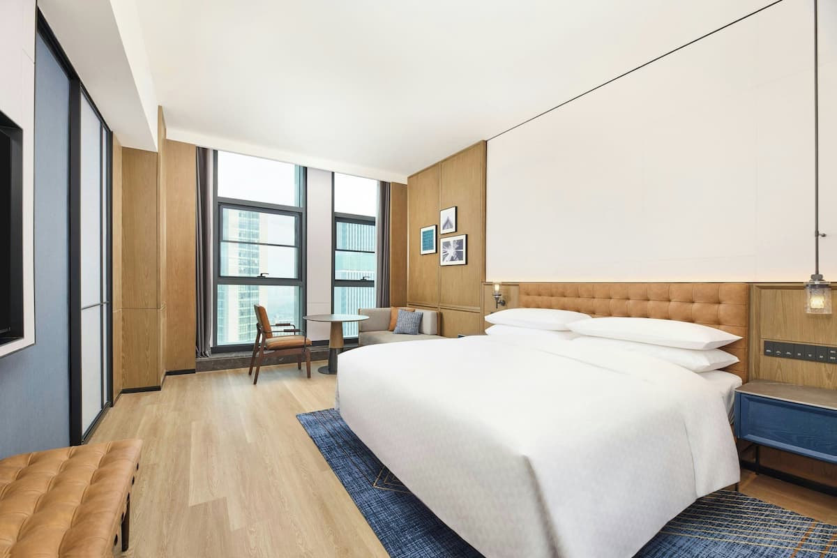 A modern hotel room with a large bed, a desk, a chair, and a view of the city through large windows.