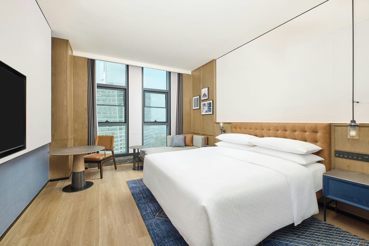 A hotel room with a bed, a desk, a chair, a TV, and large windows.