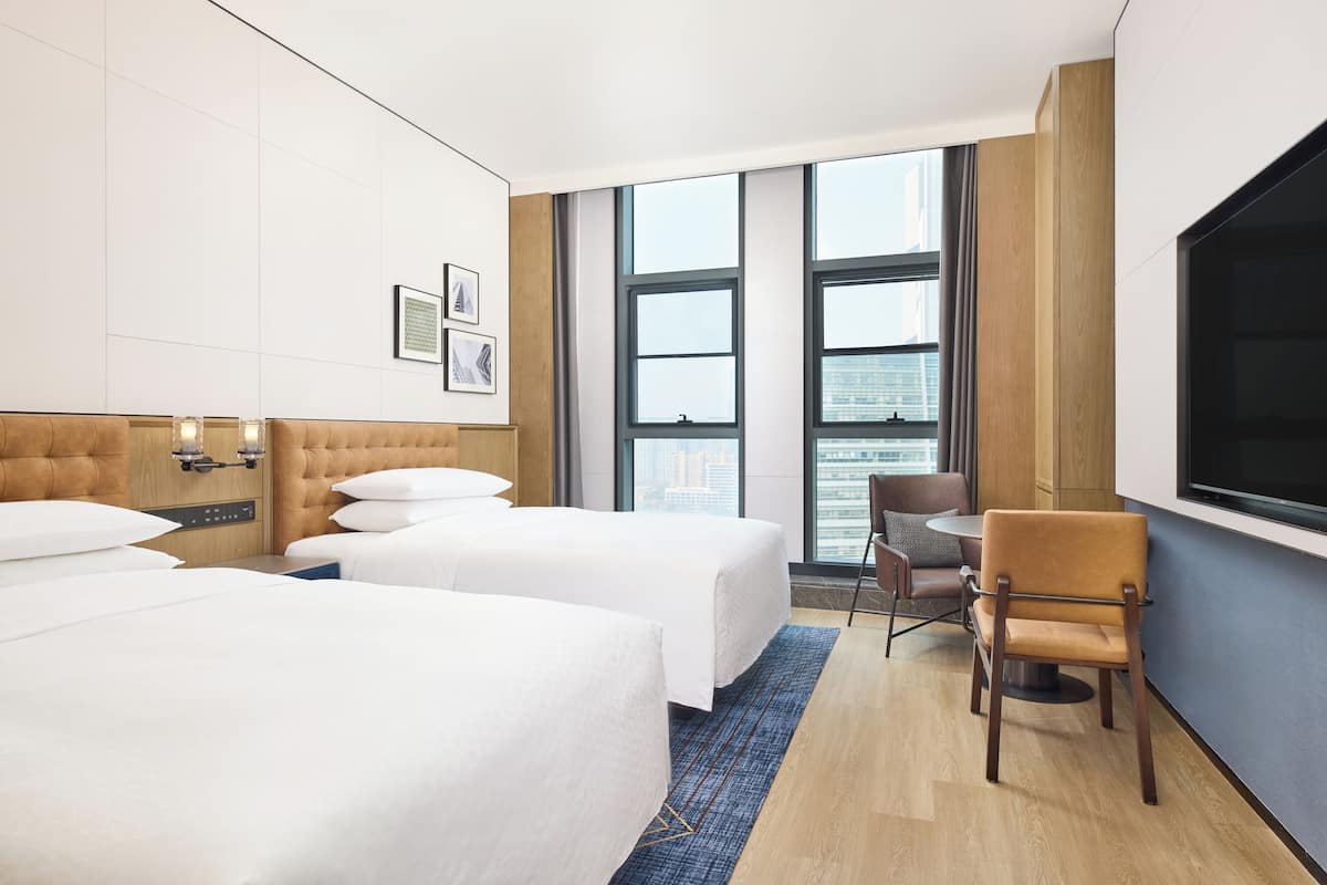 A hotel room with two beds, a TV, a chair, and a large window with a city view.