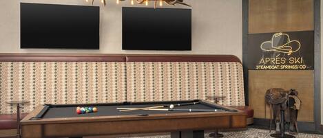 A pool table with balls, a sofa, a wall-mounted TV, and a decorative antler chandelier.
