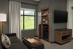 Una habitación de hotel moderna con sofá, una mesita de noche de madera, televisor de pantalla plana, microondas integrado y un refrigerador pequeño.