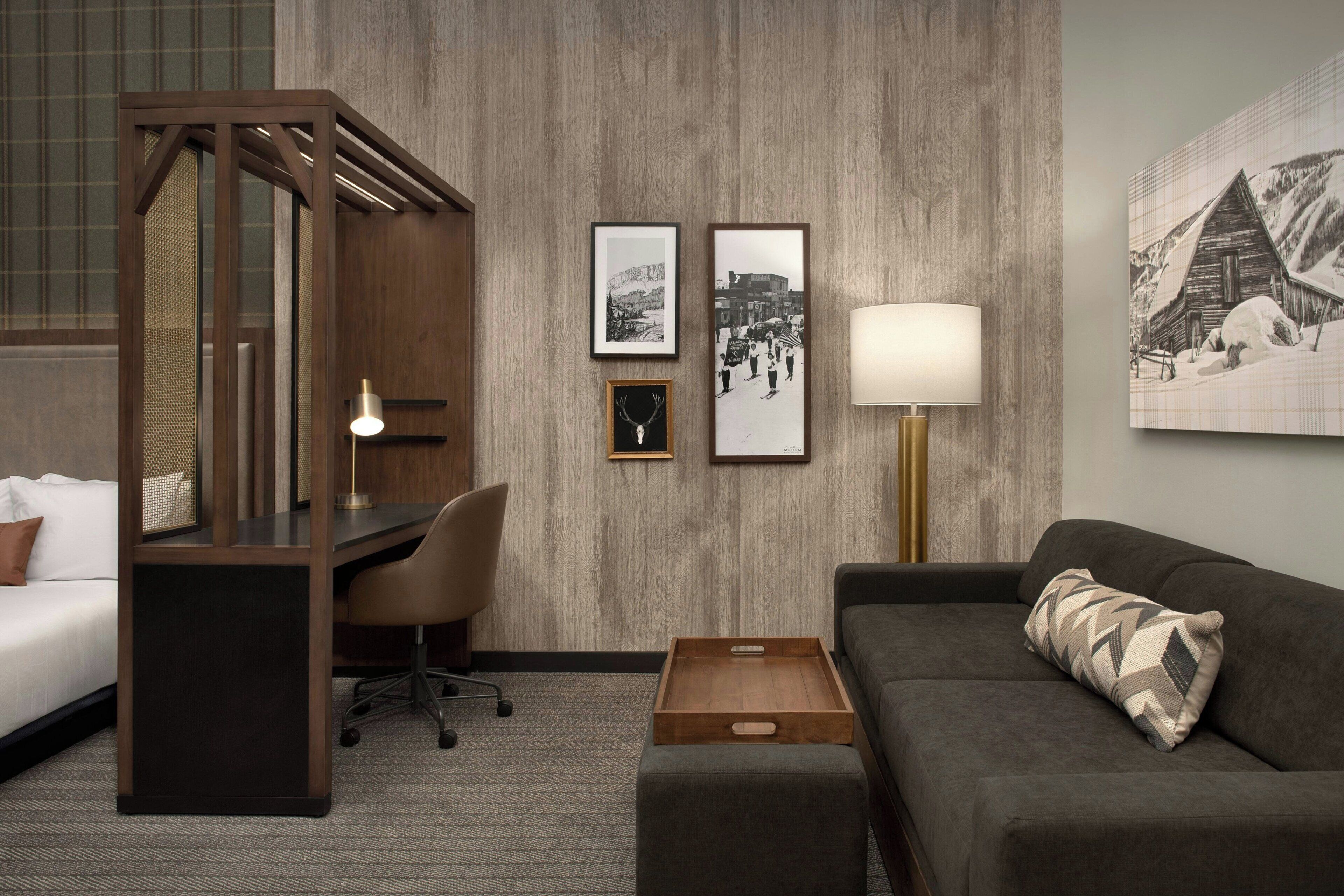 Habitación de hotel con cama, escritorio, silla y sofá. En la pared hay cuadros enmarcados y una lámpara junto al sofá.