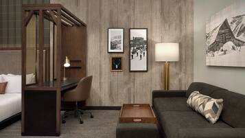 Habitación de hotel con cama, escritorio, silla y sofá. En la pared hay cuadros enmarcados y una lámpara junto al sofá.