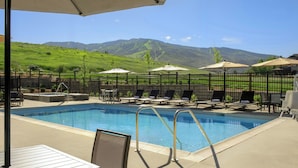 A swimming pool with lounge chairs and umbrellas, surrounded by a fence, with a view of green hills and mountains in the background. - Springhill Suites By Marriott Steamboat Springs (Steamboat Springs)