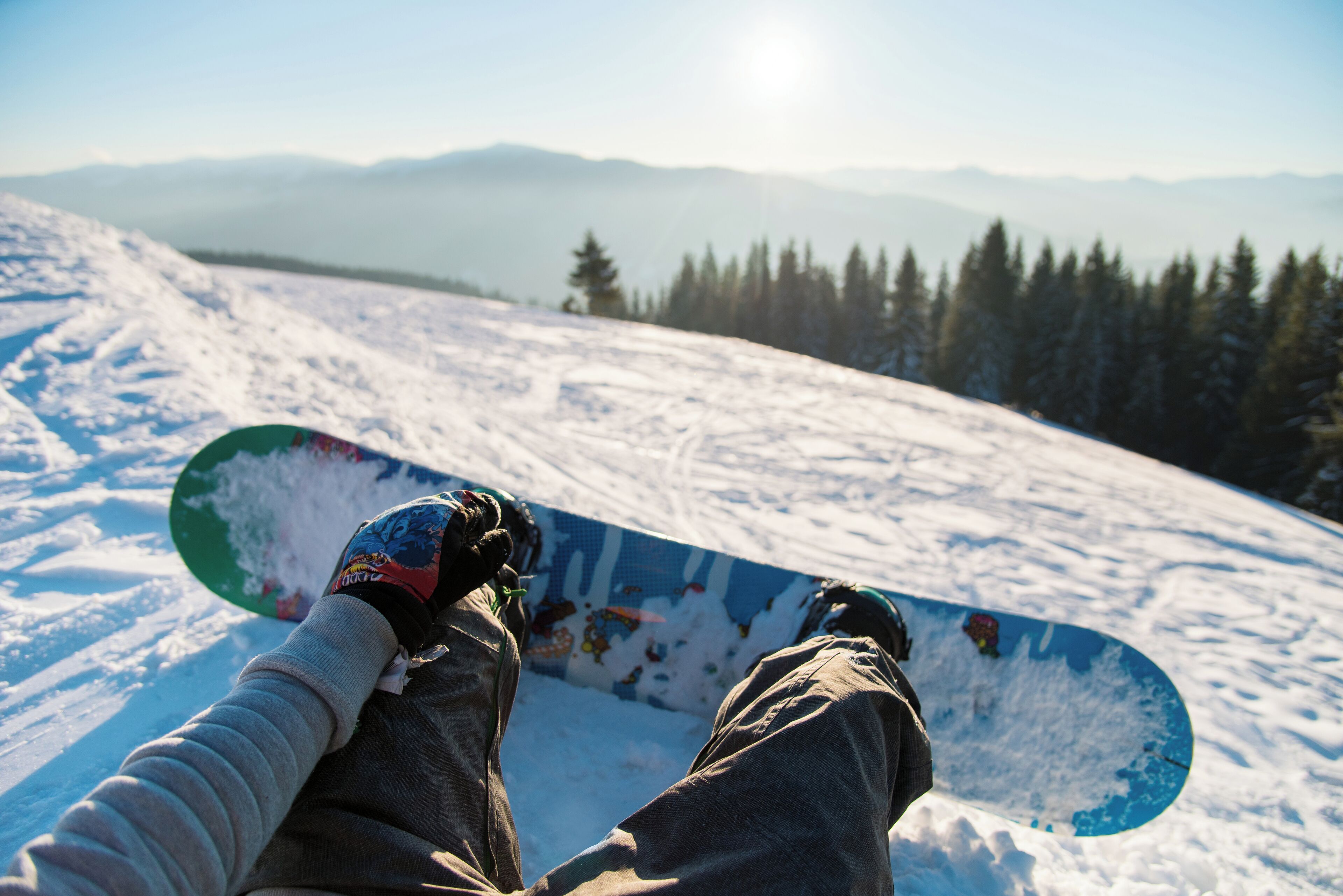 A snowboarder sitting on a snowy slope with a clear view of distant mountains and trees.