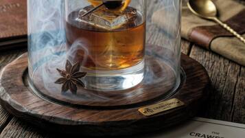 Un verre contenant un liquide ambré, garni d’une cerise et d’un zeste d’orange, placé sous un cloche de verre qui laisse échapper de la fumée, le tout posé sur un sous-verre en bois.