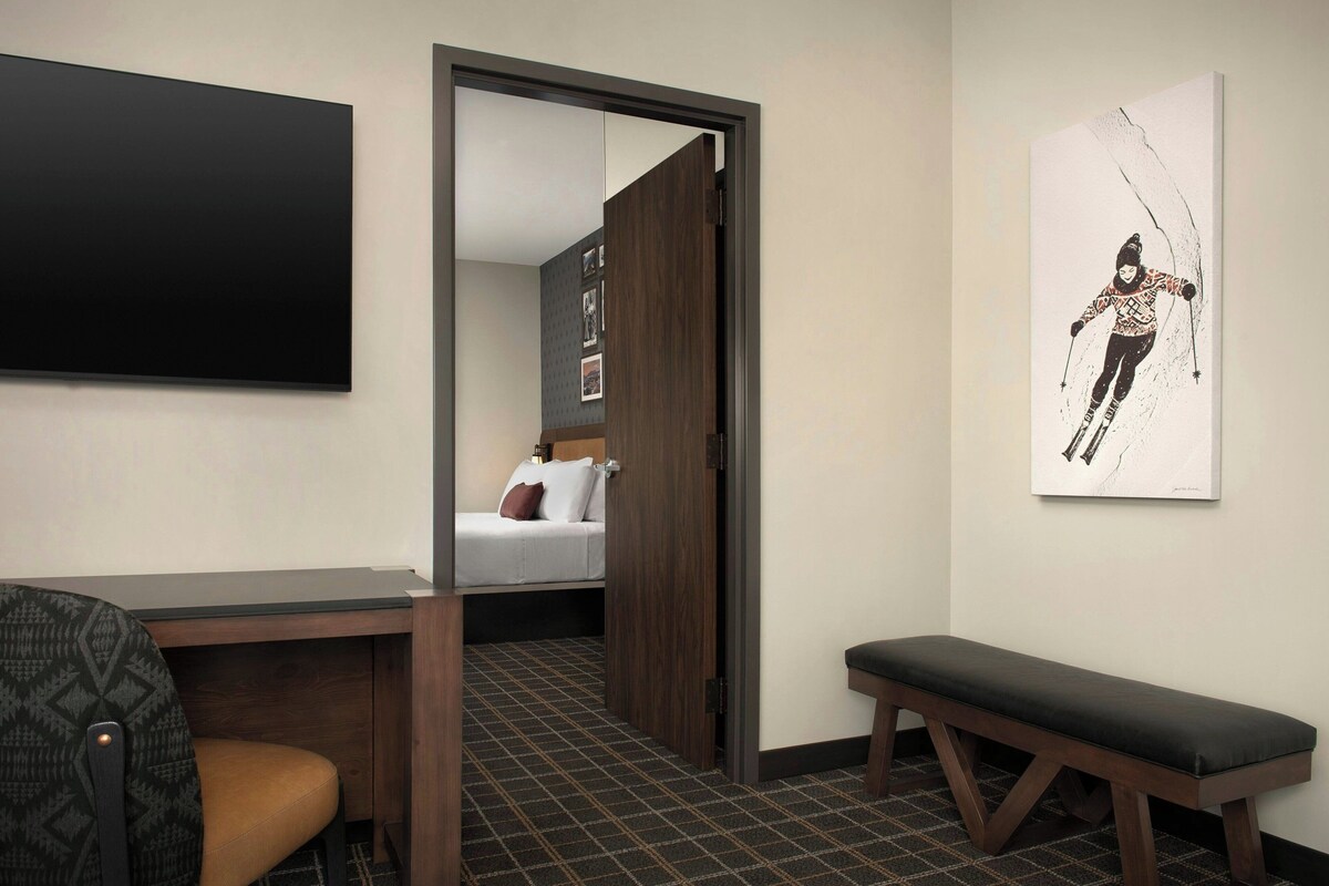 Habitación de hotel con cama, escritorio, banco, televisor y una obra de arte con temática de esquí en la pared.