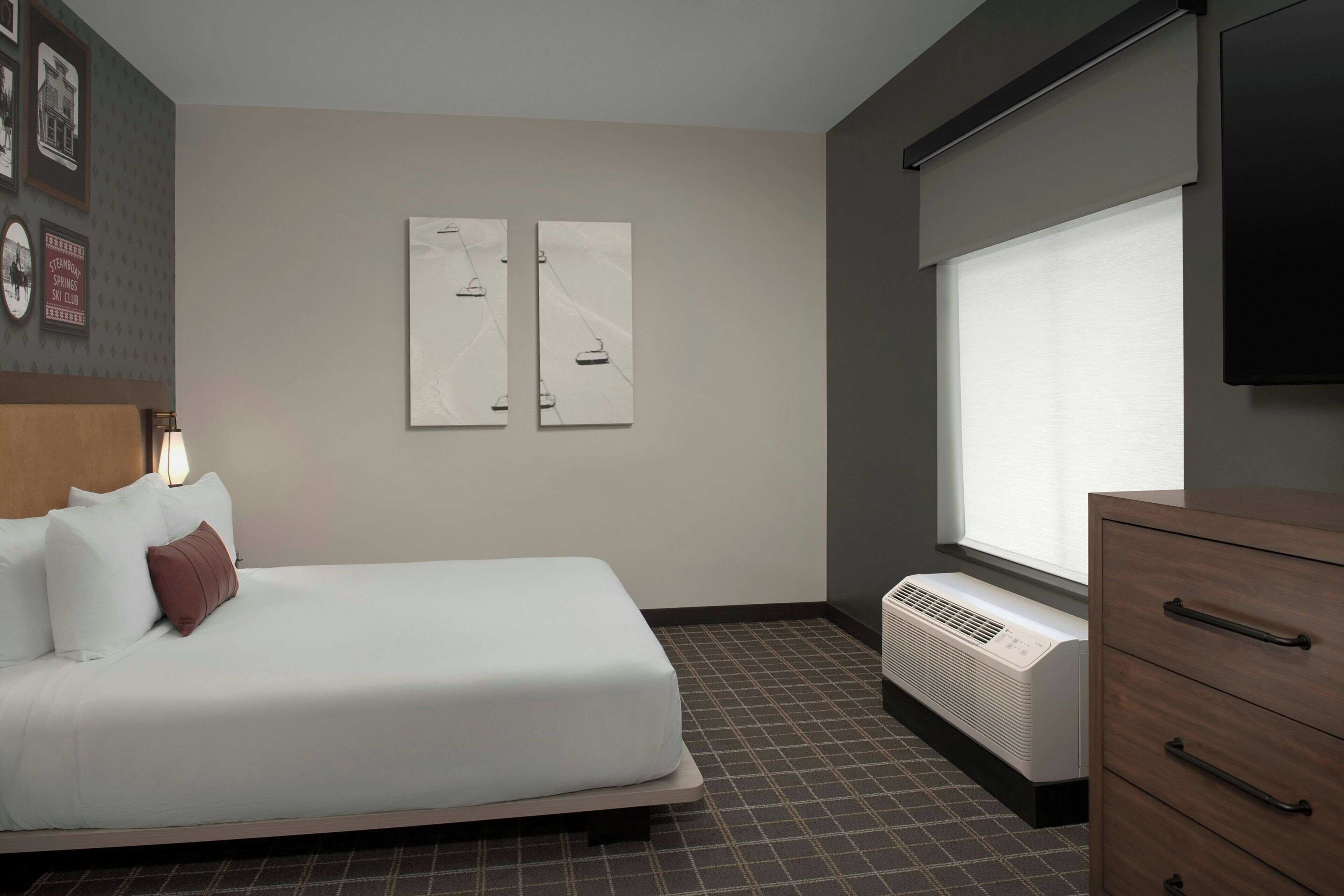 A hotel room with a bed, a wall-mounted TV, a heater, and a window with blinds.