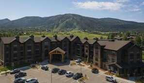 A multi-story hotel with a central entrance, parking lot, and mountainous backdrop.
