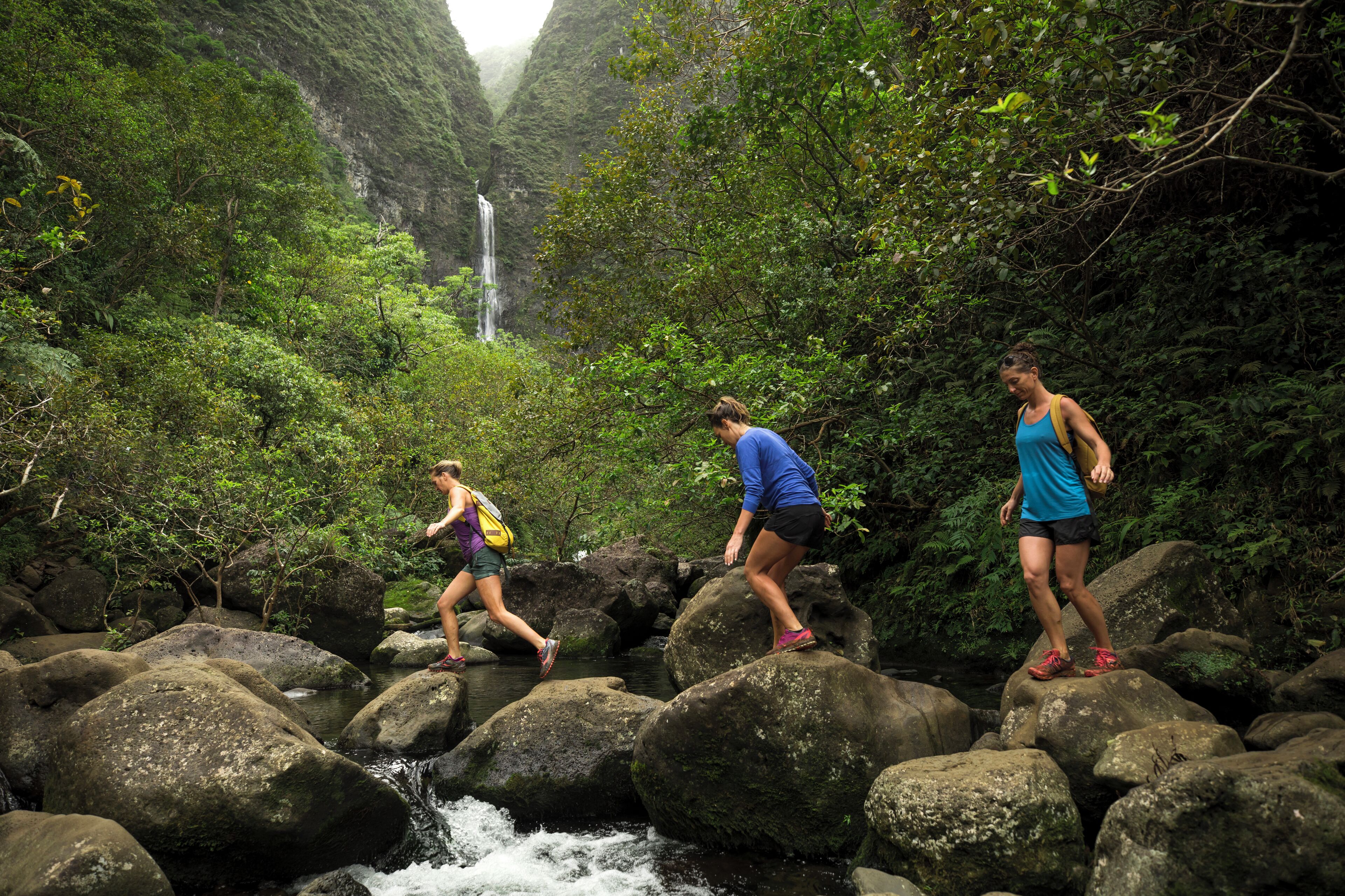Drie mensen wandelen over rotsen, met een waterval op de achtergrond.