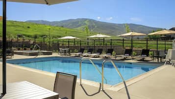 A pool with lounge chairs and umbrellas, surrounded by a fence, with a view of green hills and mountains in the background.