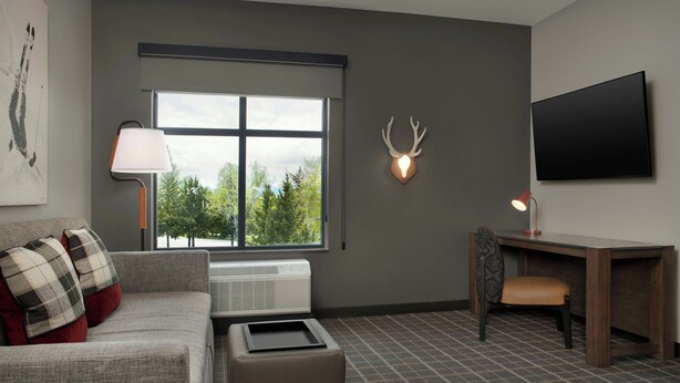 A modern living room with a grey sofa, a small ottoman, a desk with a chair, a mounted TV, and a deer head wall decoration. - Towneplace Suites By Marriott Steamboat Springs (Steamboat Springs)
