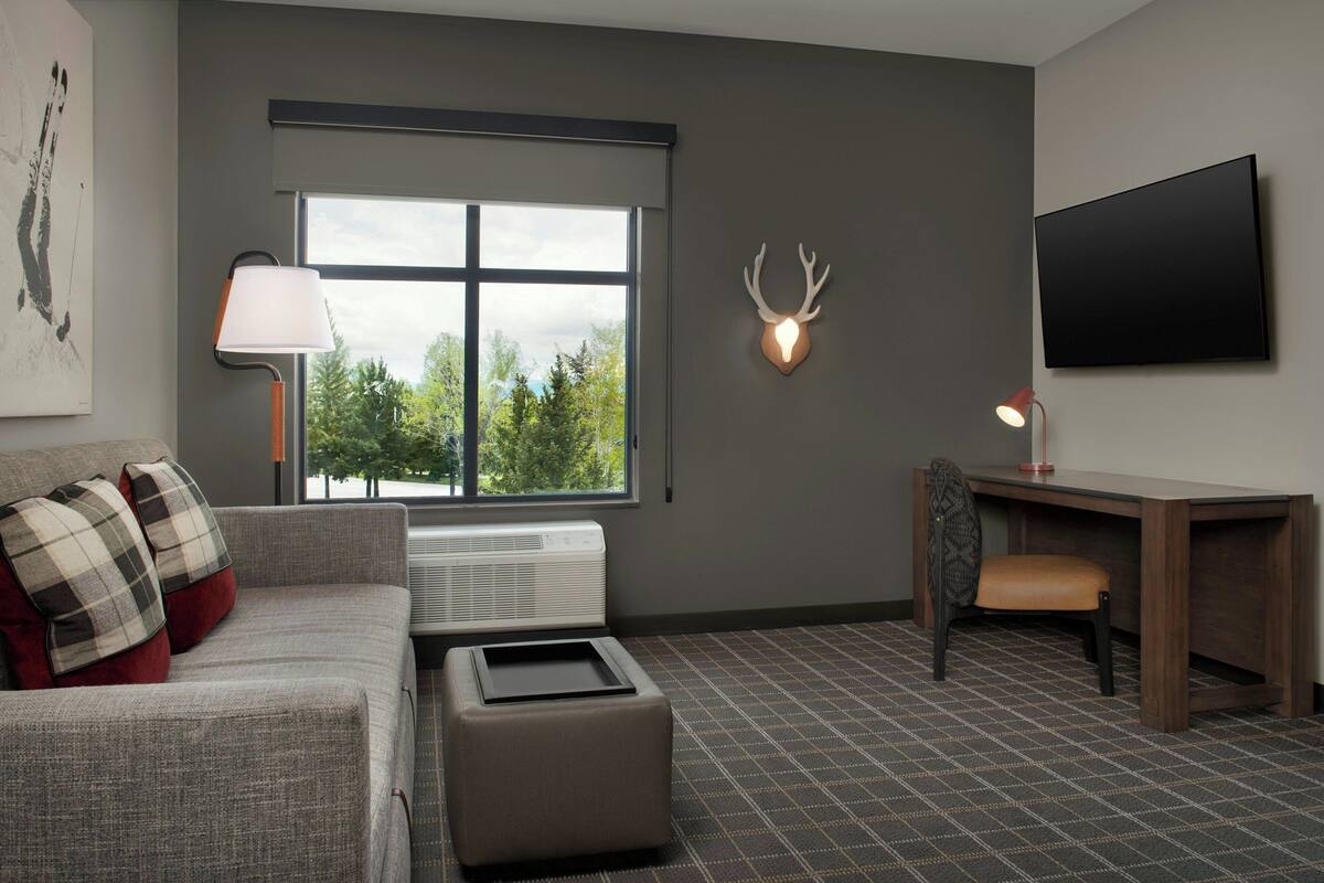 A modern living room with a grey sofa, a small ottoman, a desk with a chair, a mounted TV, and a deer head wall decoration.