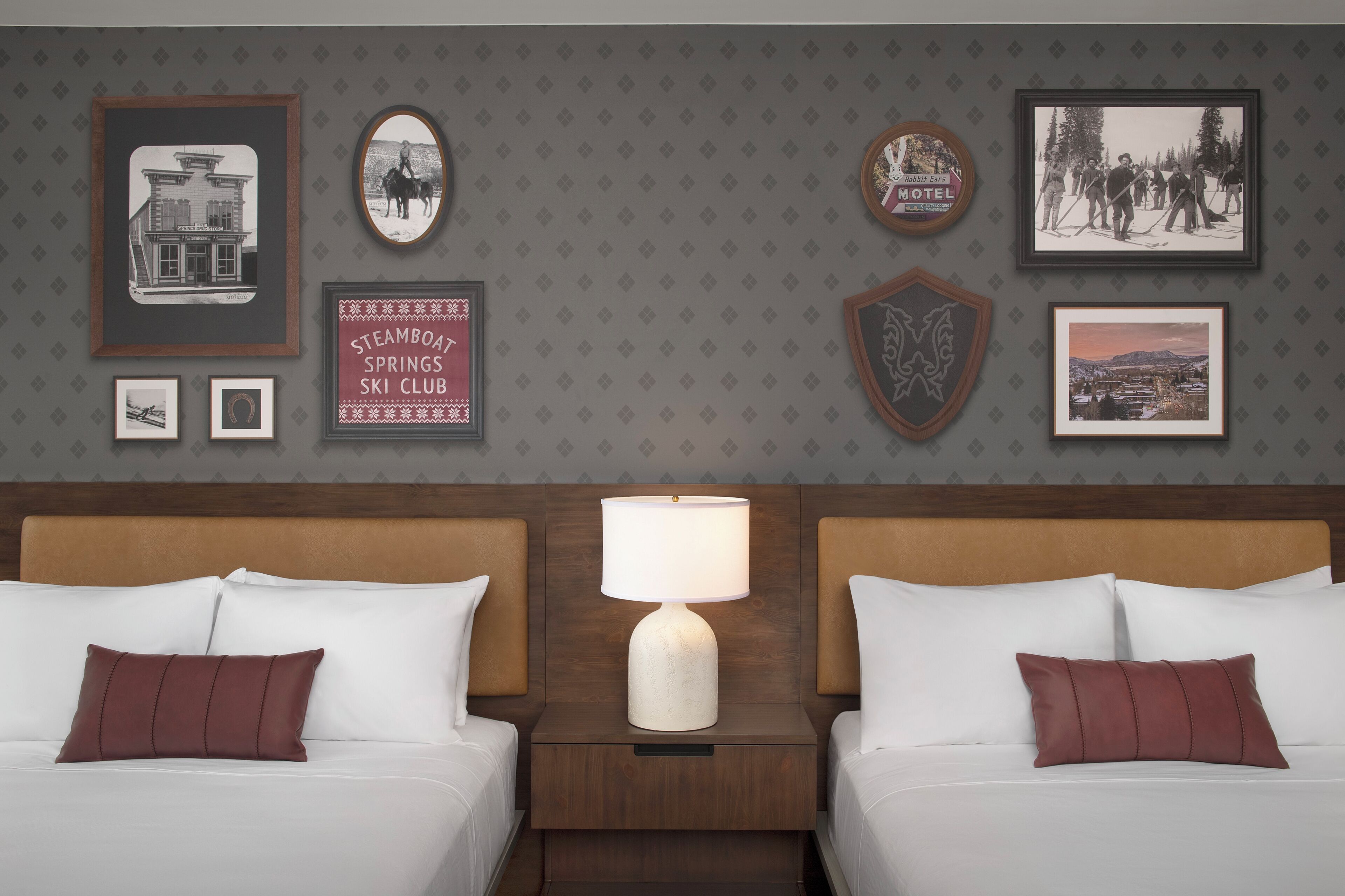 A hotel room with two beds, a nightstand with a lamp, and various framed pictures on the wall.