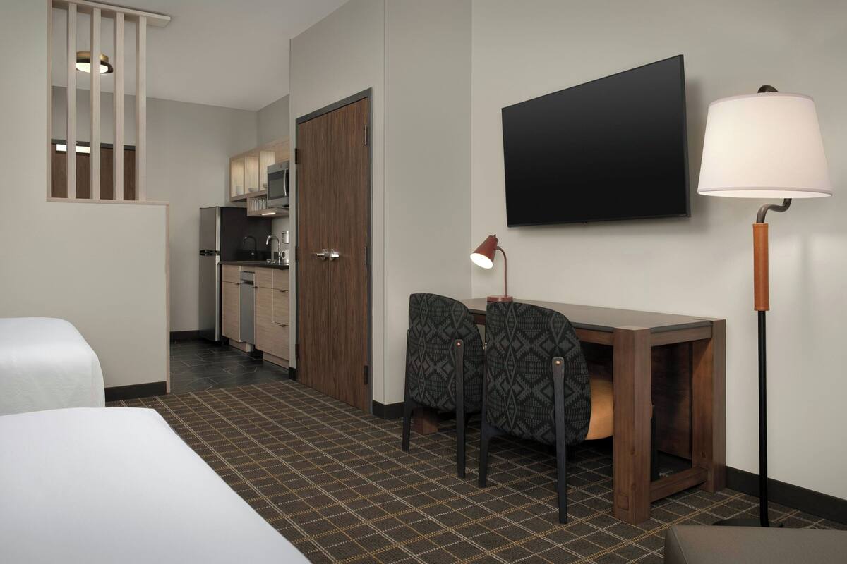 A hotel room with a bed, a desk with two chairs, a television, a lamp, and a kitchenette.