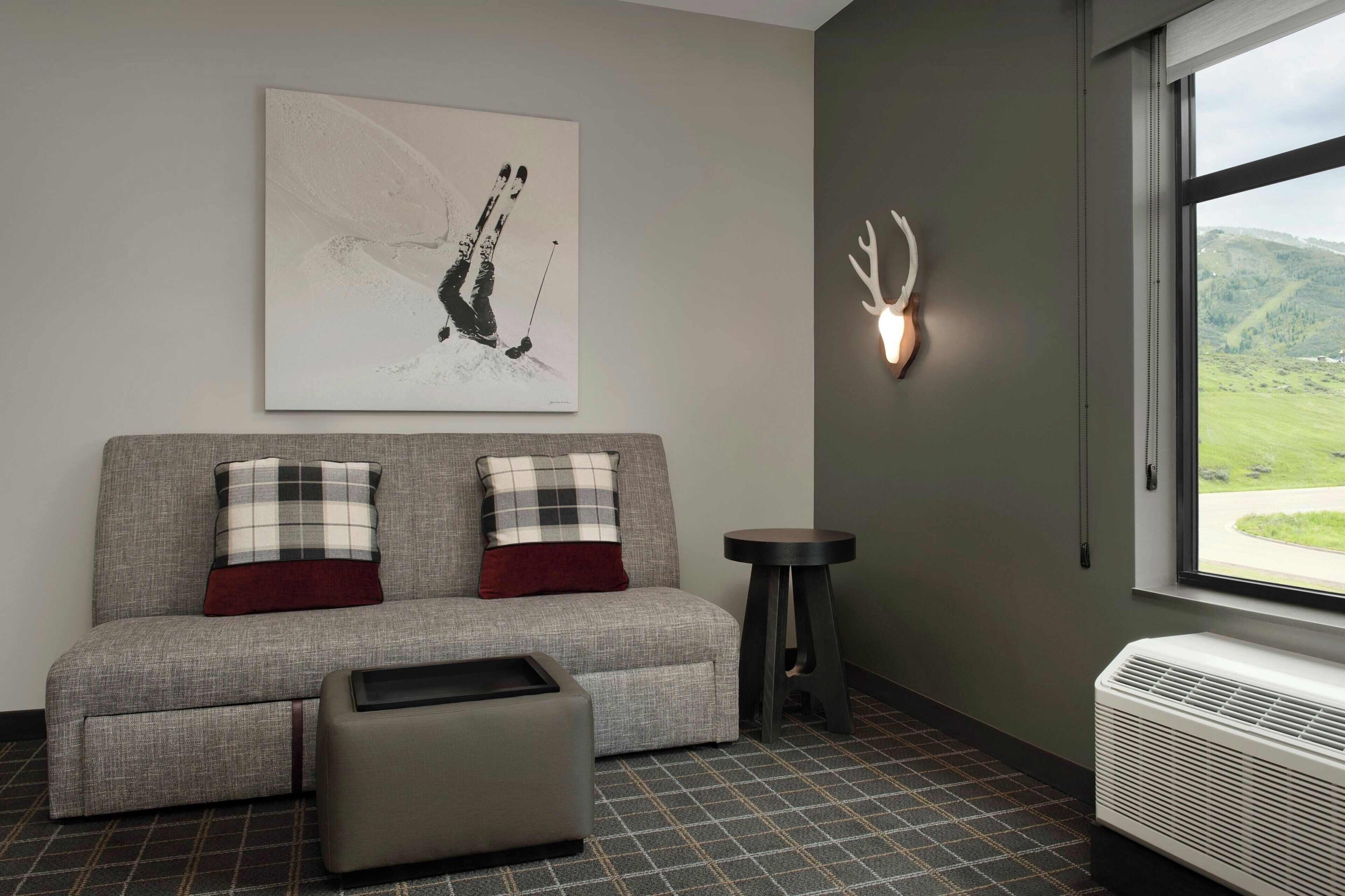 Une pièce avec un canapé gris, deux coussins à carreaux, une petite table, un lampadaire fixé au mur et une fenêtre donnant sur des collines verdoyantes.