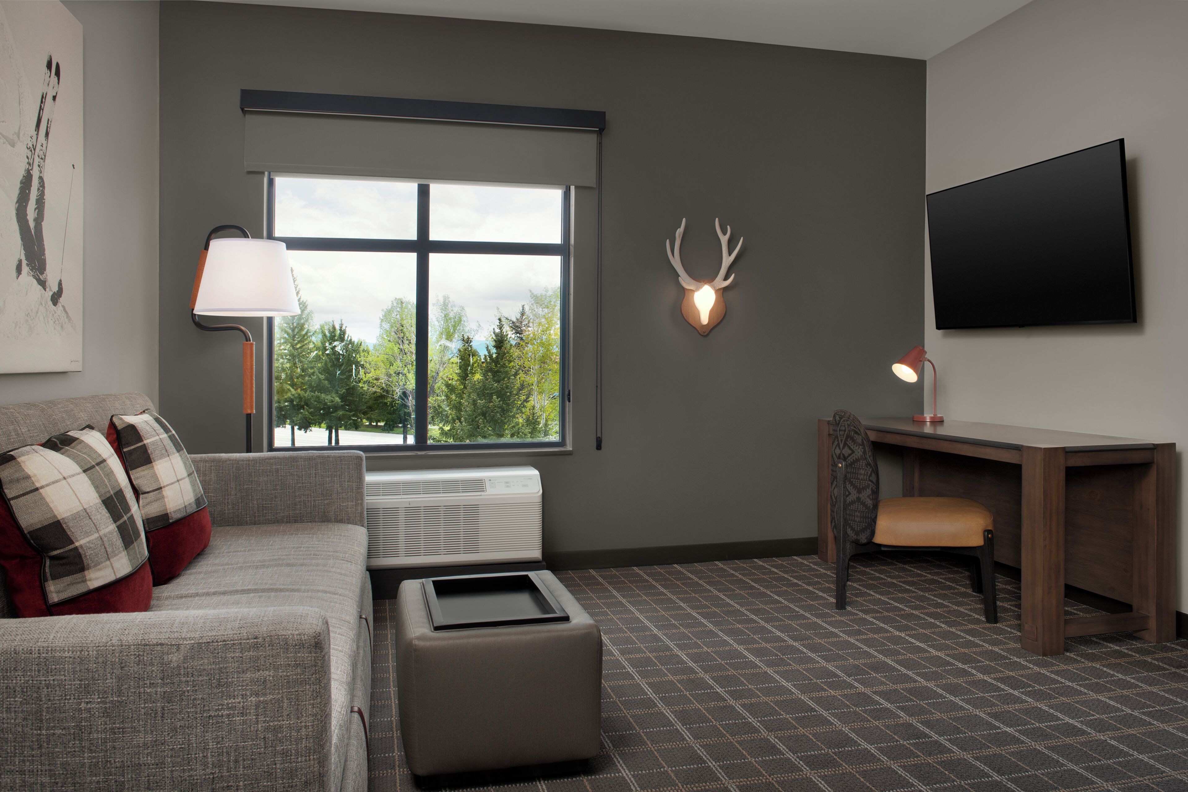 Una sala de estar moderna con un sofá gris, un pequeño reposapiés, un escritorio con una silla, un televisor montado en la pared y una decoración de cabeza de ciervo.