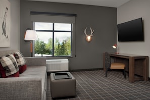 A modern living room with a grey sofa, a small ottoman, a desk with a chair, a mounted TV, and a deer head wall decoration. - Towneplace Suites By Marriott Steamboat Springs (Steamboat Springs)