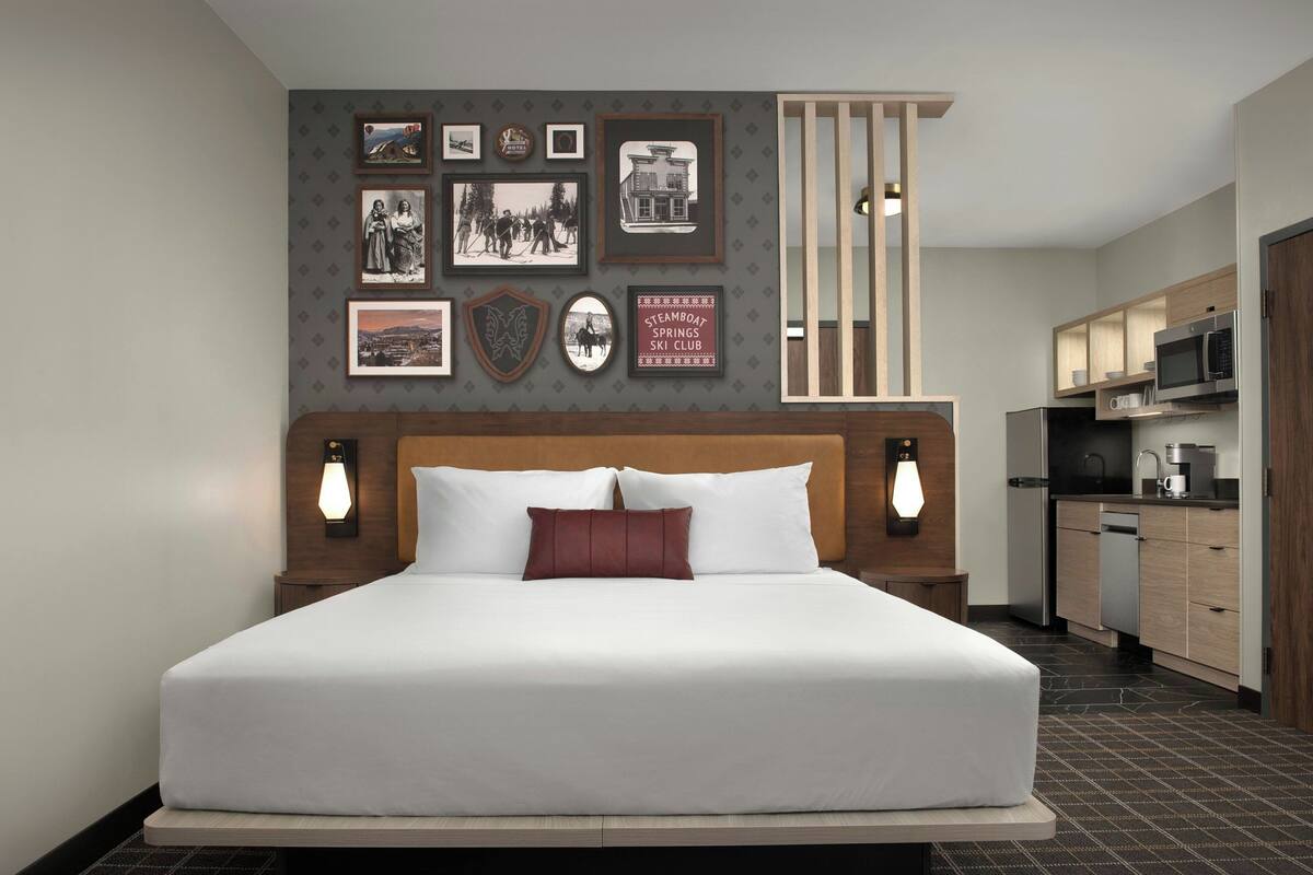 A hotel room with a large bed, a wooden headboard, a wall with framed pictures, a kitchen area with modern appliances, and a patterned floor.