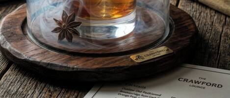 A glass of whiskey with a cherry and an orange peel garnish, served under a glass dome with smoke, on a coaster with a star anise, at The Crawford Lounge.