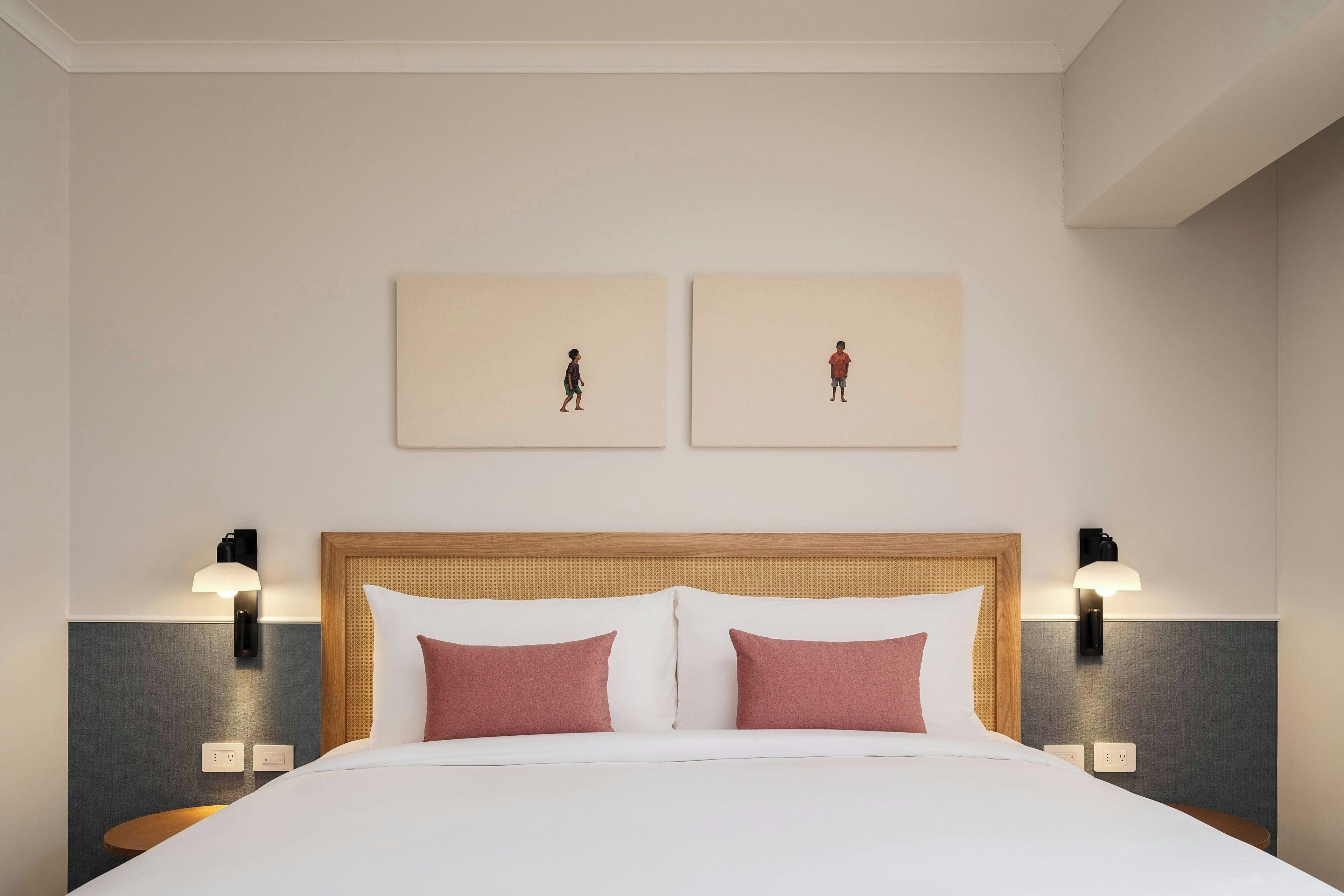 A hotel room with a bed, two wall-mounted light fixtures, and two framed artworks on the wall.