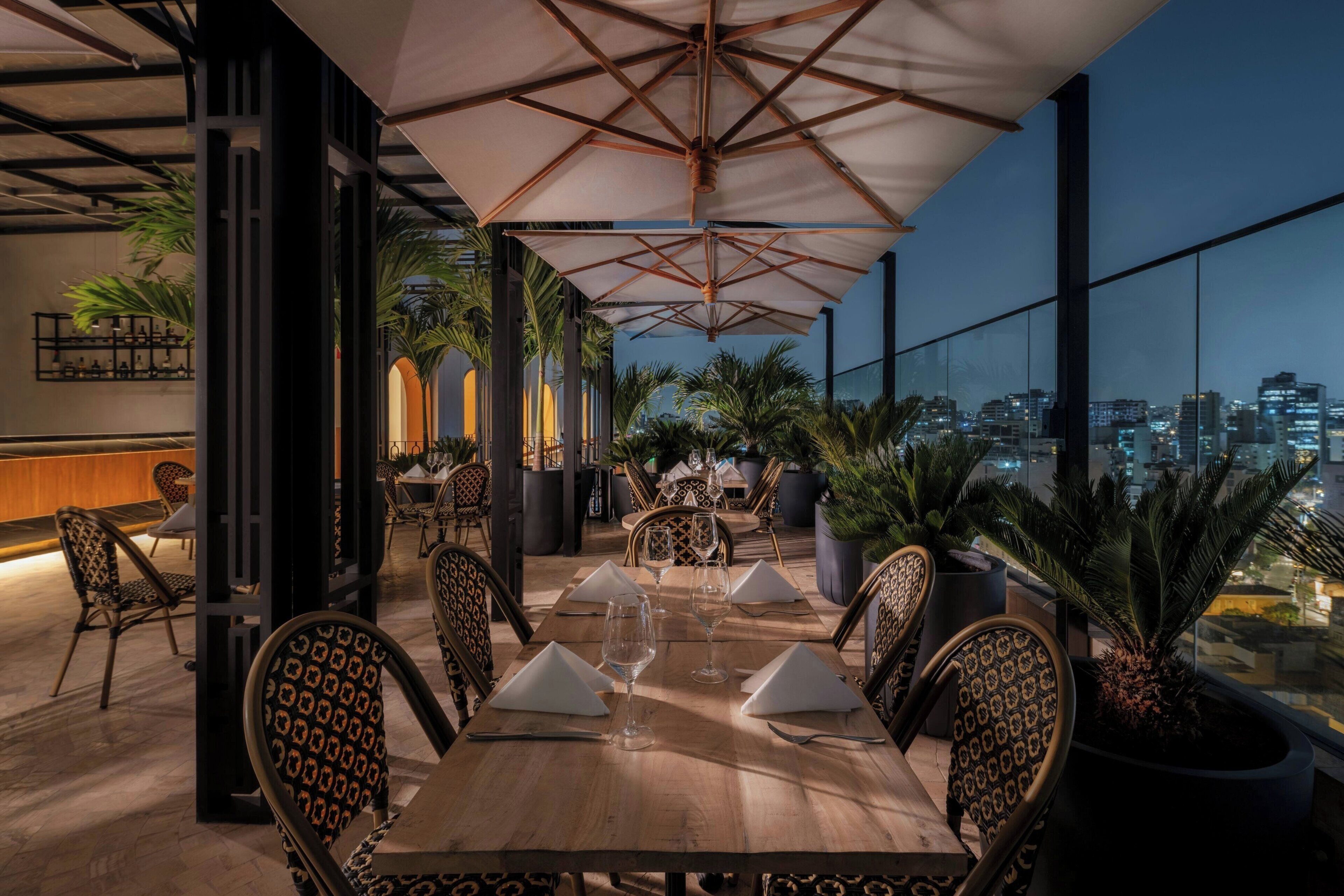 Terraza comedor con vista panorámica de la ciudad, mesas dispuestas con servilletas blancas y copas de vino, y sillas estampadas.