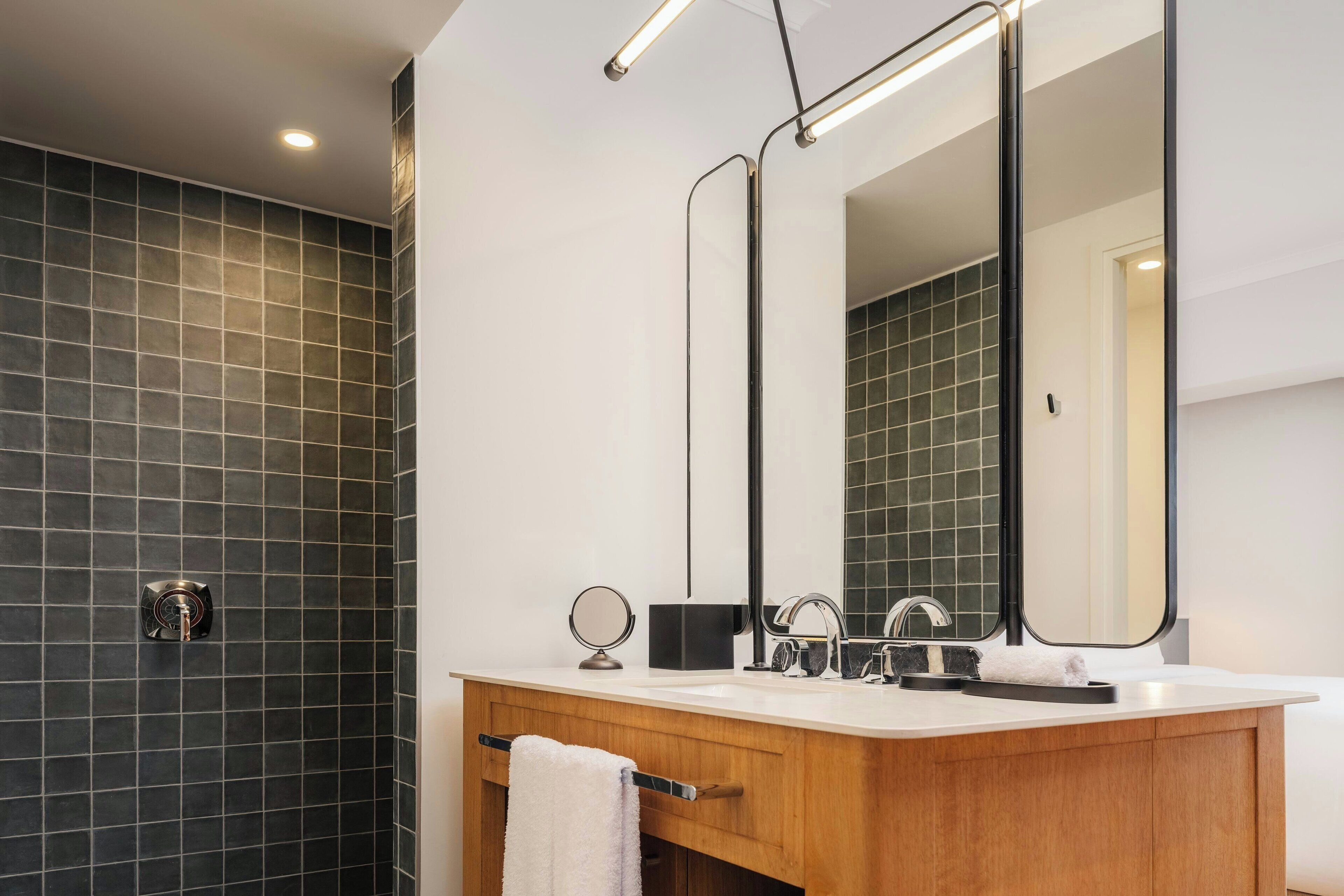 Une salle de bain moderne avec un mur recouvert de carreaux foncés, un meuble-lavabo en bois, un miroir et un porte-serviettes.