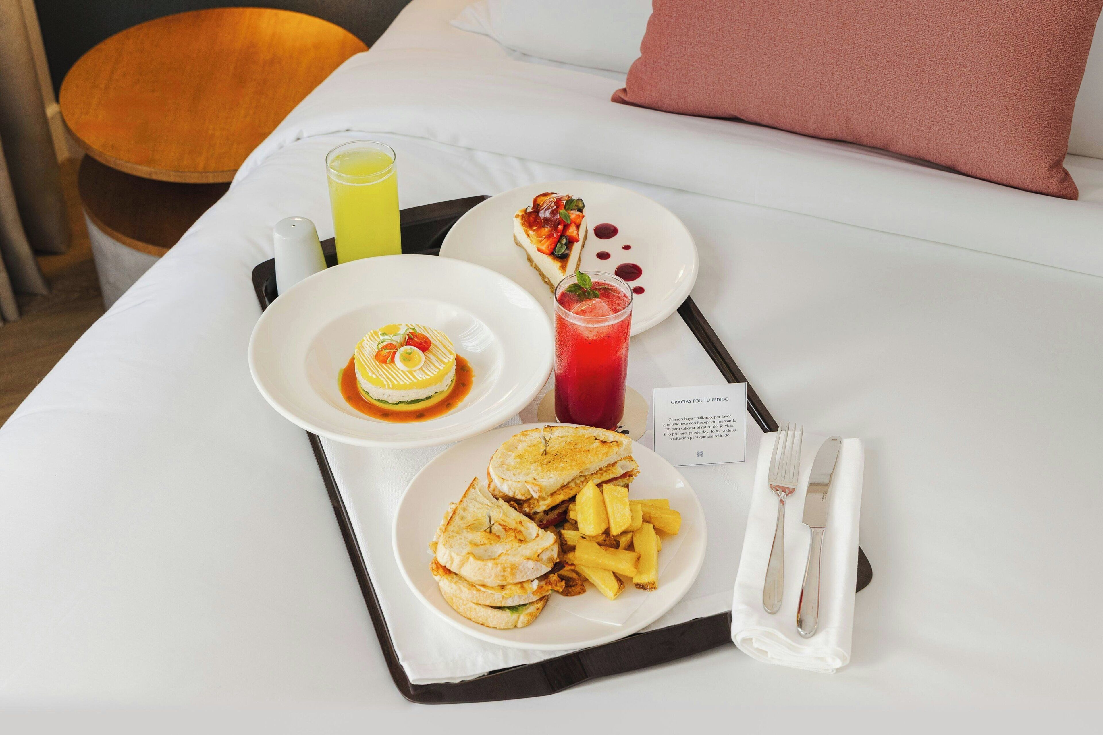 Una bandeja de desayuno bien ordenada sobre una cama, con una almohada, un plato de comida, un vaso de jugo y un postre.