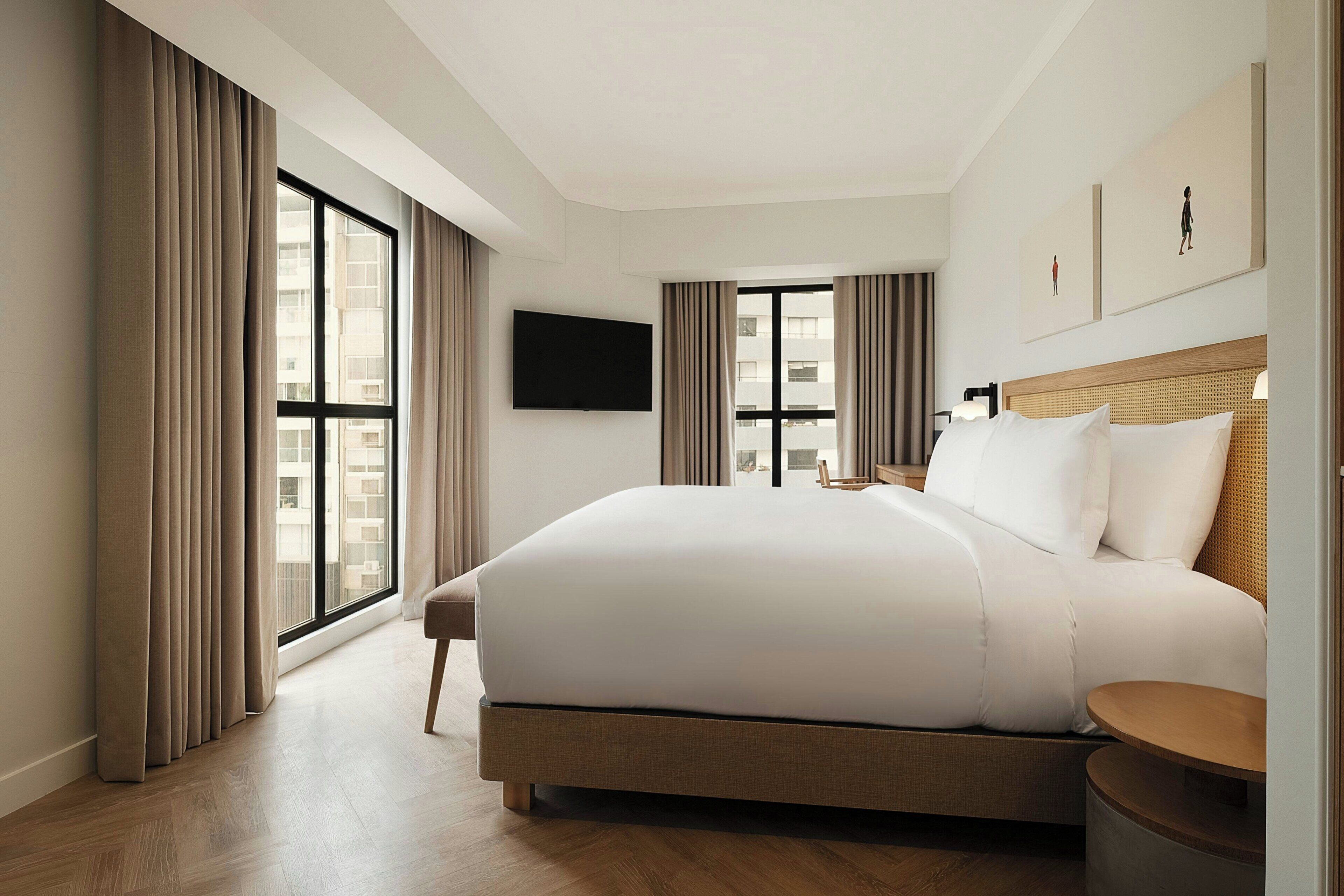 A hotel room with a large bed, a flat-screen TV, and a view of the city through the windows.