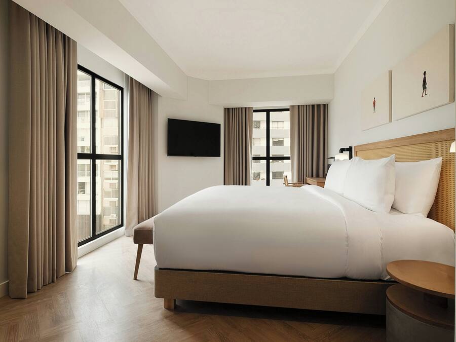 A hotel room with a large bed, a flat-screen TV, and a view of the city through the windows.