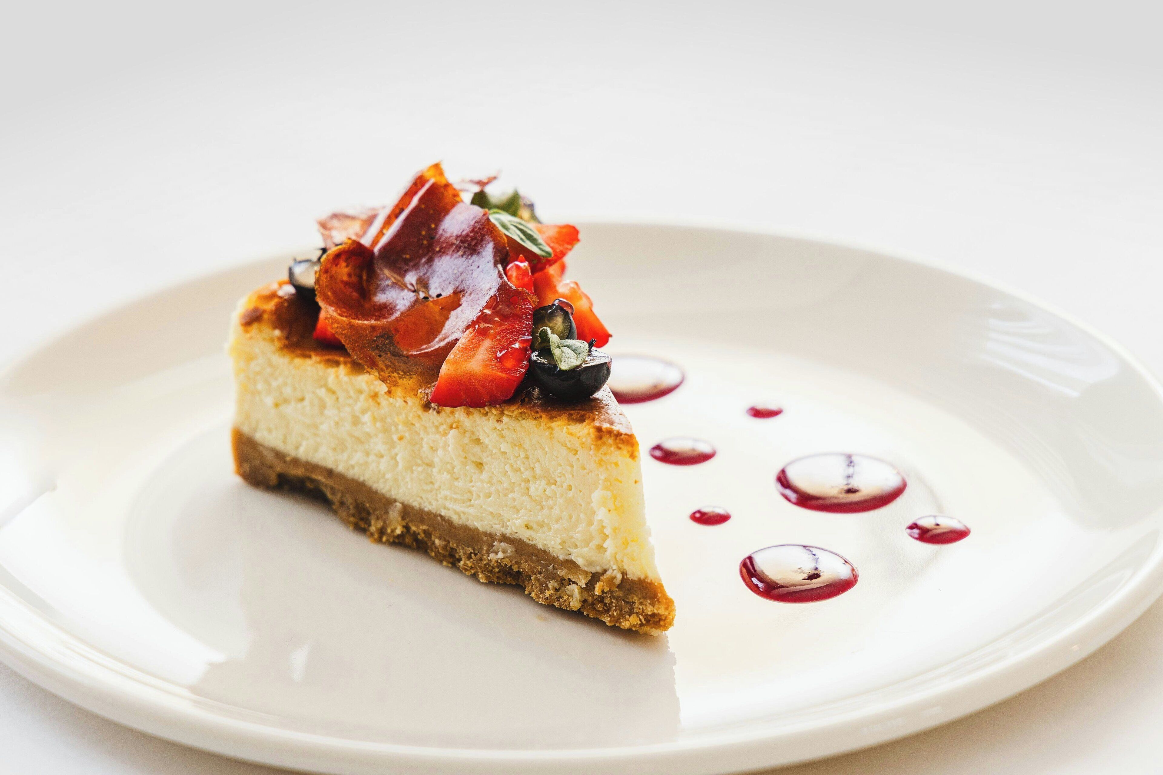 Ein Stück Käsekuchen mit karamellisierter Oberfläche, verziert mit roten Früchten und Kräutern, serviert auf einem weißen Teller mit vereinzelten Soßtröpfchen.